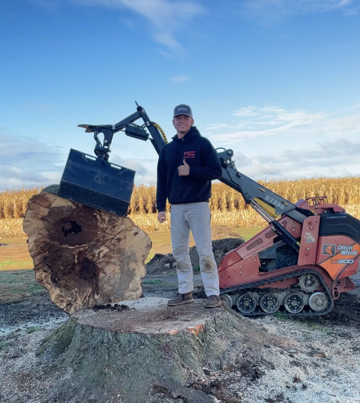 Better Way Land Management LLC crew member standing on large tree stump with Ditch Witch SK800 mini track loader in rural field setting