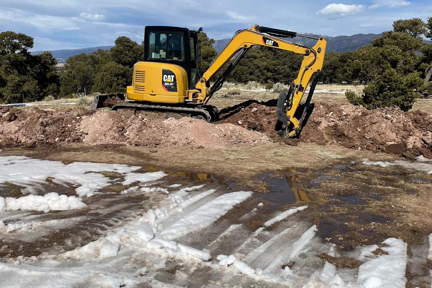 Better Way Land Management LLC CAT 305.5E mini excavator digging trench through rocky soil at rural construction site with mountains visible.