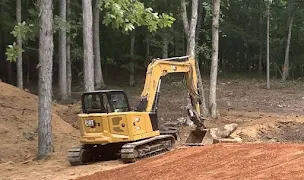Better Way Land Management LLC CAT mini excavator clearing wooded terrain and moving earth at forested construction site.