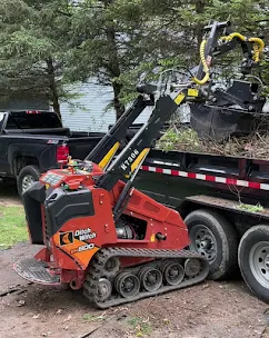 Better Way Land Management LLC Ditch Witch SK3000 compact track loader with grapple attachment loading onto flatbed trailer at wooded job site